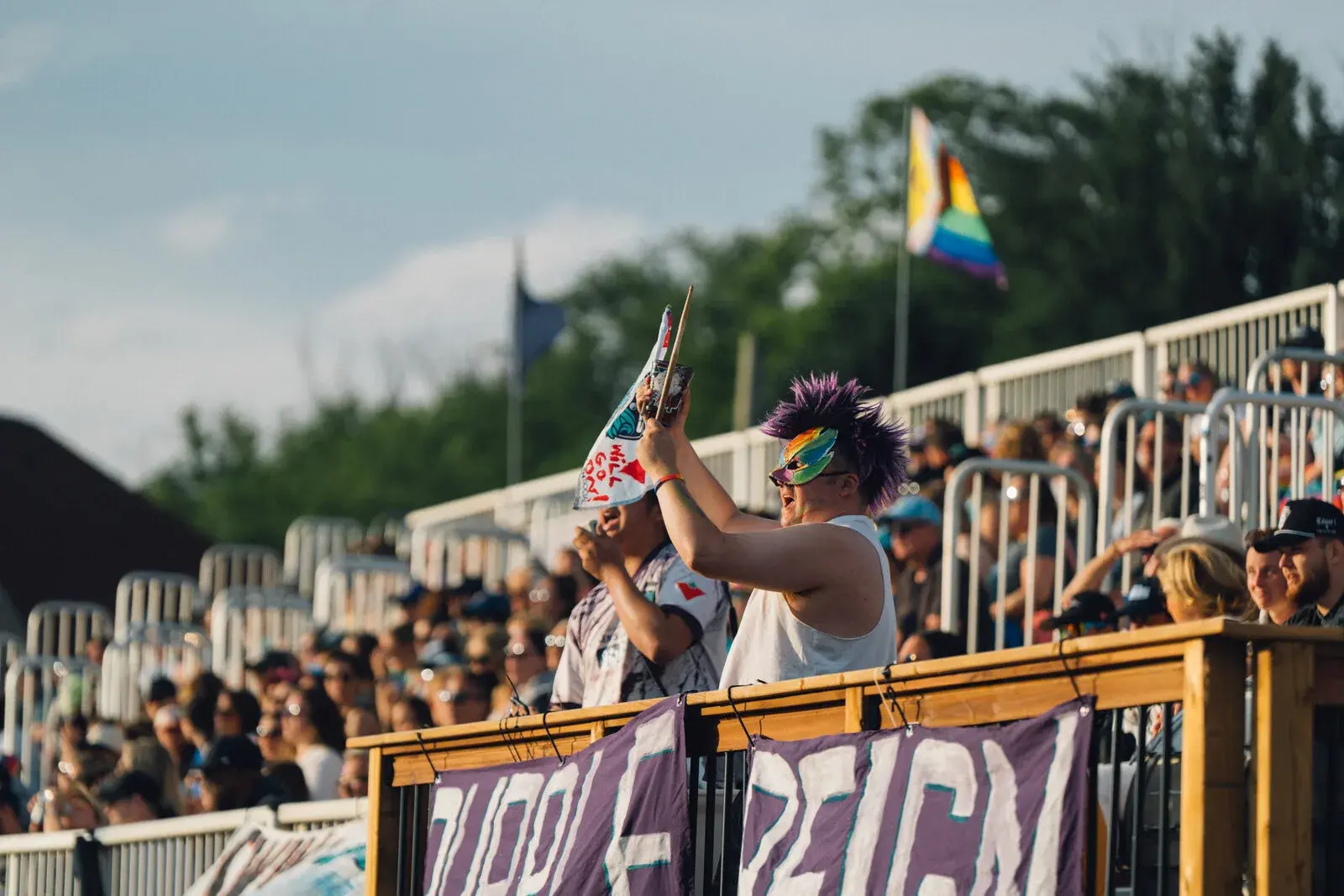 Fans in the stands at a summer event, one wearing a rainbow mask and waving a flag
