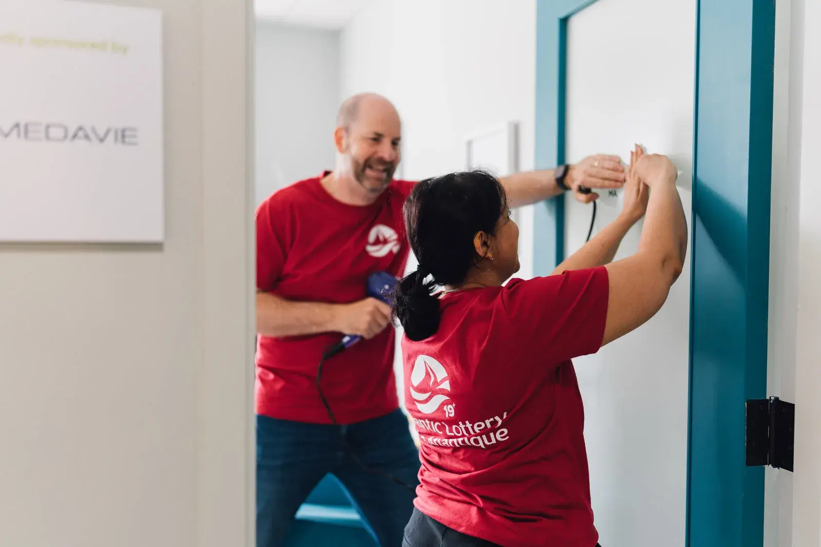 Two Atlantic Lottery employee volunteers in red shirts mounting a sign on a wall